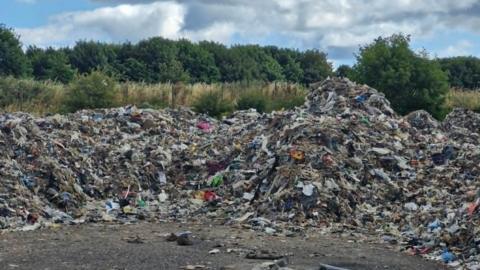 Mounds of rubbish, which looks like a mixture of shredded plastic and dirt, on the floor. Behind it are hedges and trees