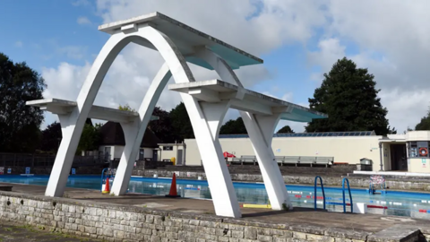 large 1930s white diving board with three boards in front of the outdoor pool in Stroud. Pool has blue steps and lane floats. 