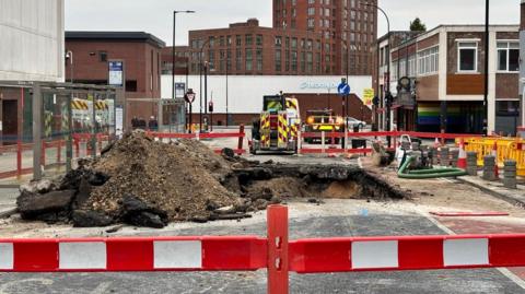 A construction site on street. There is a pile debris in the middle of the street which is cordoned off by red and white barriers. Several construction vehicles and buildings are in the background.  