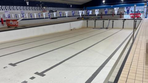 An empty six-lane, 25m (82ft) swimming pool with a sloped white lining, with black lines on the base to denote where the lanes are. There are builders' materials around it and plastic orange safety fencing. There are beige non-slip tiles around the walkways surrounding the pool.