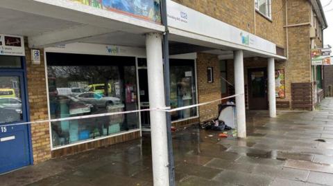 A shopping precinct is in the image, and there is tape outside of a building which is a pharmacy. There is a fridge outside of the building stood on the floor with fire damage.
