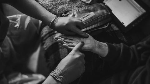 Black and white image of a a pair of hands. One is wearing gloves and the other has a plaster being placed on her palm. There is medical kit in the background.