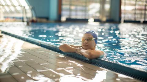 A young swimmer wearing yellow and blue goggles and a blue swimming cap is resting her chin on her arms, which are on the side of a swimming pool. She is in the pool, looking into the distance.