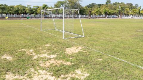 A white football net on a pitch with large patches of bald and dry grass. A fence runs along the edge of the pitch in the background with trees behind.