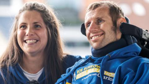 Lindsey Burrow, who has long, dark brown hair and is wearing a blue hooded top. She is smiling and kneeling down next to Rob Burrow who is in a wheelchair which has a neck support. He is smiling and is also wearing a blue hooded top with yellow writing on which says 'Rob Burrow Leeds Marathon'.