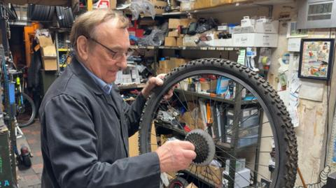 A man standing in a workshop while holding a bike wheel with a chunky black tyre. He has short brown hair and wears black-rimmed glasses, a grey fleece and a blue shirt. The workshop is filled with shelves and boxes containing tools and equipment. A bike and more tyres can be seen in the background.