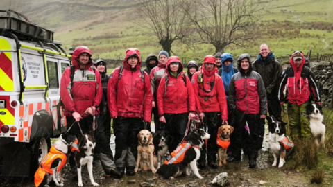 A group of mountain rescue volunteers stand next to a vehicle with a number of rescue dogs on leads. They are all very wet. 