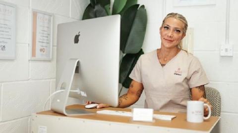 A woman with blonde hair tied in a ponytail. She is wearing a beige top and is sitting at a desk. She is sitting in front of a Mac desktop computer with a white mug near her hand.