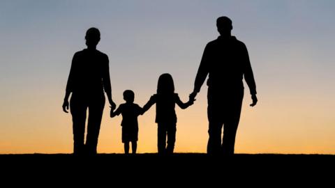 A man and woman holding hands with their son and daughter. They are silhouetted in the photo with the sunset behind them.