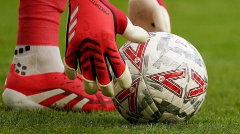 A close up of a football on grass. A goalkeeper wearing gloves leans down to pick the ball up.