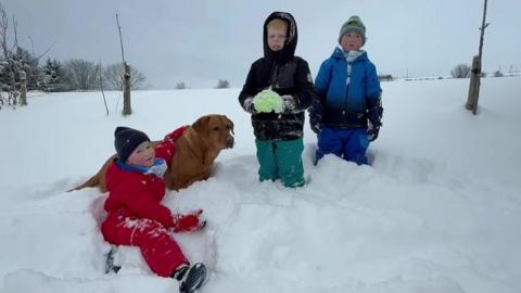 Three children and a dog are in the snow. One child is sat in a red snow suit with their arm around the Labrador. The other two children are standing wrapped up in coats, hats and gloves. The snow comes up to their knees