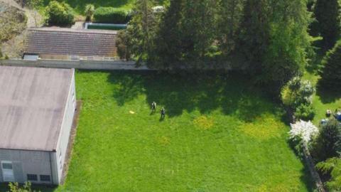 Shot from above, two men stand in a large green open space with a flat roofed building on the left of the site and what appears to be houses, partly screen by mature conifer trees, at the top of the image.
