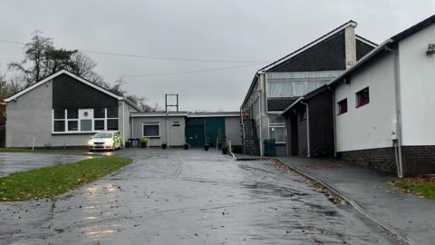 A derelict grey and white building. Looking from the entrance out front. A police car parked outside with headlights turned on. A patch of green grass just in front. Wet and rainy weather. 