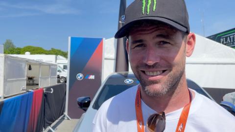 Sir Mark smiling and wearing a black Monster Energy cap, white t-shirt with sunglasses hooked onto the neckline, and an orange TT lanyard. There are tents and barriers behind him on a sunny day.