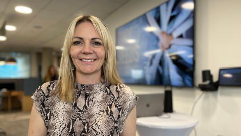 Chloe Fossé smiles at the camera. She has blonde hair and brown eyes, and is wearing a sleeveless print dress. Behind her to the right is a large TV screen attached to a wall with the Tiny Seeds name on and people putting hands into the centre of a circle.