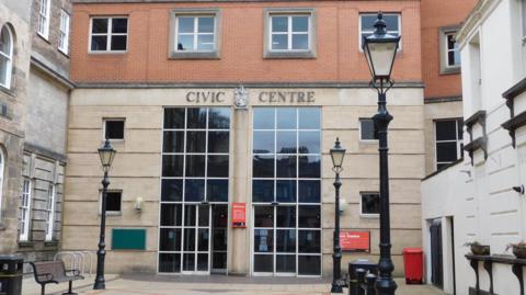 An entrance to a building that is made from light stone at the bottom and red brick at the top. Two panels of rectangular windows make up part of the building's entrance. There are three traditional, black lampposts at the front of the image.