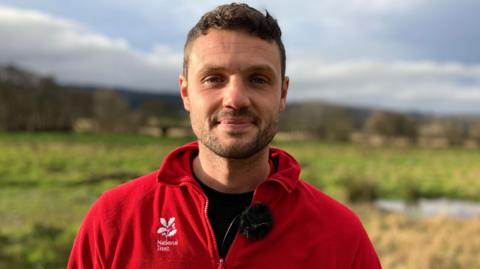 Jack Siviter. He is pictured outdoors in Holnicote Estate. The landscape is out of focus but green fields and trees can be seen behind him. Jack has short dark hair and facial hair. He is wearing a red fleece with a white National Trust logo on the chest. He is looking directly at the camera and smiling 