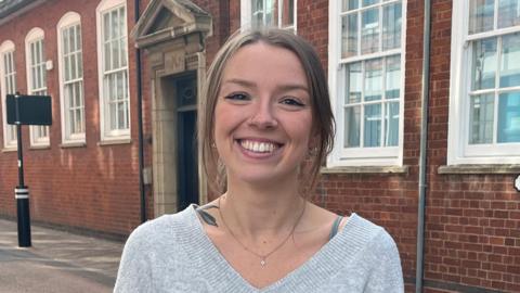 Olivia Dieterich, a white woman in her twenties standing in Guildhall Lane in Leicester. She is wearing a grey sweater and has her hair tied up. She is smiling. 