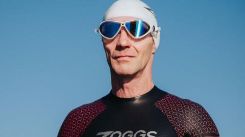 A close up of Martin, he wears a swimming cap and goggles and looks out into the distance, he wears a black wetsuit.