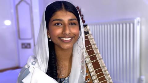 A woman with a white scarf over her head holding a dilruba. 