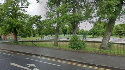 Trees in a park with grass and park furniture. There is a brick hut. There is a green fence and a road with road markings.