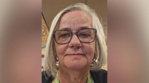 A close-up portrait of Helen Edwards with shoulder-length grey hair wearing dark-framed glasses, photographed indoors against a softly lit background.