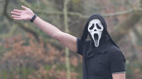 A man wearing a mask from the horror film Scream, raising his right arm in a wave. He has a black polo shirt on.