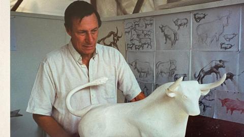 Sculptor Laurence Broderick standing behind the maquette the Bull with previous sketches behind him 
