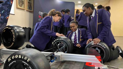 Children in purple school uniform carrying out a mock pit stop on a frame with racing car wheels attached.