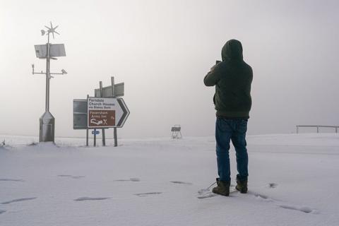A man dressed in a dark jacket, facing away from the camera, takes a picture of snow on a sign - the ground is covered by snow and the sky is grey or white.
