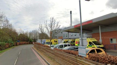 The outside of Hereford's County Hospital. The image is taken from the road side outside the A&E department. The hospital's façade is made up of red brick and grey cladding panels. A small roof with pillars extends out over the car park. Several yellow ambulances are parked beneath it with at least four cars parked in the car park facing away from the hospital.