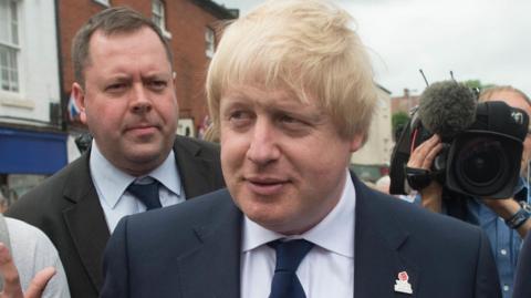 A crowd of people standing around former UK prime minister Boris Johnson. Johnson is wearing a navy blue suit and navy blue tie with a poppy badge. Behind him is Will Walden, who is wearing a black suit and tie. There is also a TV camera operator.