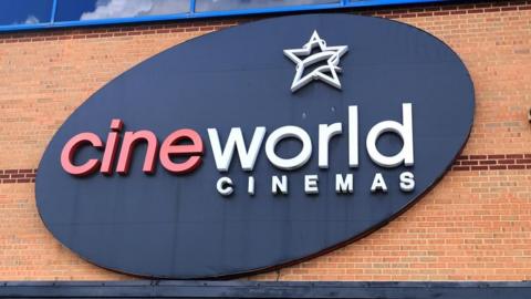 A large oval black Cineworld Cinemas sign on a brick wall. The "Cine" is in red and the rest of the letter are in white.