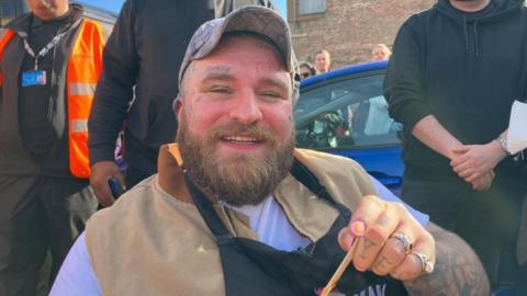 A heavily tattooed man with a beard and wearing a cap sitting down on a chair in front of a blue car with people stood behind him. He is wearing a black apron over a camel-coloured top and is smiling at the camera. He is holding wooden cutlery in one hand.