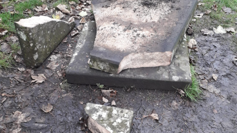 A gravestone on a patch of muddy ground is seen broken into a number of pieces.