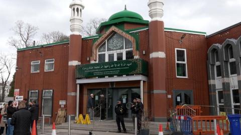 A wide shot of the Manchester Central Mosque. It is a red brick building with a green dome on top. There are multiple members of the police standing outside.