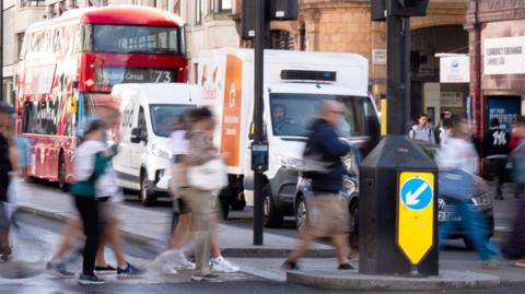 A view of Oxford Street shows two large white vans, a 73 double decker bus to Oxford Circus and a car with pedestrians crossing at traffic lights