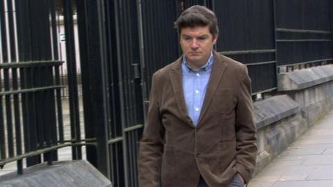 Michael McMonagle with brown hair wearing a blue and white checked shirt walks along side iron railings