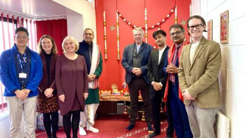 A group of people standing in front of a red shrine area in the new Hindu temple and smiling at the camera. There are red and white varamala flower garlands hanging from the wall, and lots of red white and gold religious symbols and objects sitting on the shrine. 