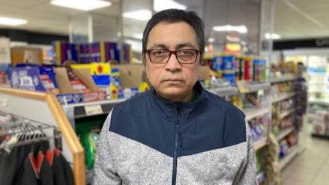 A man wearing a blue and grey zipped jumper with dark hair and glasses is standing in a convenience story with items on the shelves behind him.