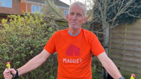 Pete Turfrey wearing a bright orange t-shirt with the silhouette of a house above the word 'Maggie's'. He is standing in a garden beside bushes and a wooden fence, holding two Nordic walking poles on either side of him. He has white hair, blue eyes, and is smiling at the camera. On the inside of his left forearm there is a tattoo of Pauline's name. 