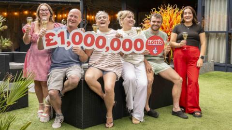 A family-of-six holding a cut out reading £1,000,000. The family consists of a mother and father, three adult daughters and an adult son. They are all smiling and holding glasses of champagne.