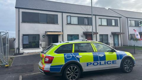 A row of houses with a police car sitting out the front of them. One house has damage to a front door following an arson attack.