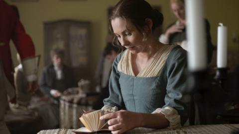 Actor portraying Jane Austen is sat at a table flicking through a book.