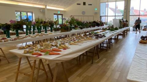 A picture of the room where the exhibitions are taking place. There is a number of tables with plants, fruits and vegetables on display. It is indoors and there is a wooden floor below the tables.