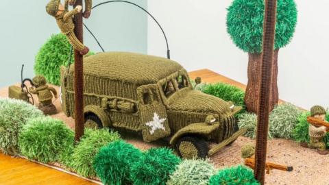 A green crocheted army vehicle and five crocheted little green soldiers. They are in a knitted scene with trees and bushes. 