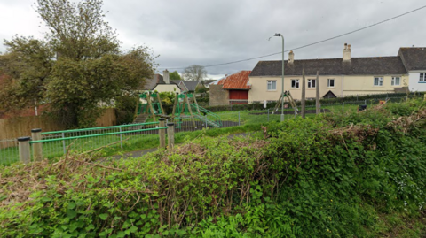 A picture of the play park in Kingsteignton. It is a large park with an area for children to play. It is surrounded by metal fences and hedges around the area.