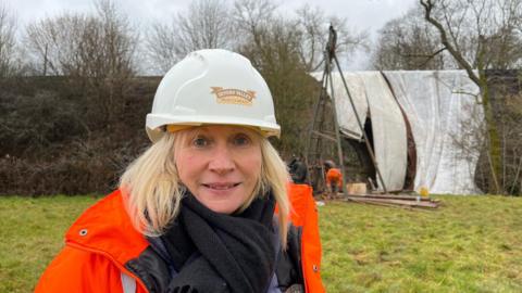 Lesley pictured in front of the borehole with the covered-up bridge in the background. She's wearing a white hard hat with "Severn Valley Railway" written on it and an orange high-visibility jacket over a black jumper and black scarf. She is standing in a green field with some trees at the edge. The sky is grey and overcast.