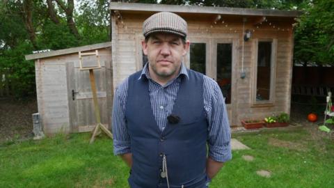Peter Chapman, wearing a waistcoat and flat cap. A wooden summer house can be seen behind him at the end of a patch of grass.