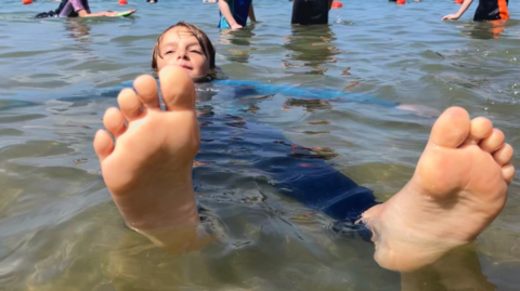 A boy wearing a wetsuit floating on his back in the sea pool. The soles of his feet are close to the camera.